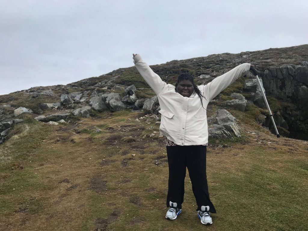 In a full-body shot against a background of stony cliffs and late-season grass, Charlene raises her arms in celebration after climbing a large hill. 