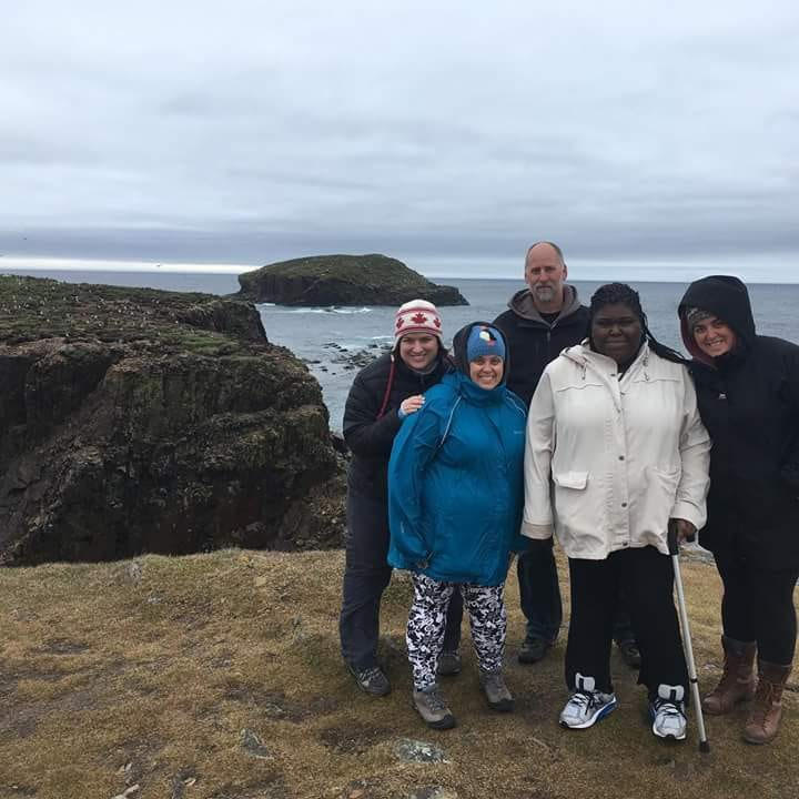 Against a backdrop of craggy cliffs and grey ocean, Charlene stands with four YACCer friends to the right of the frame. They are dressed for cold weather. 