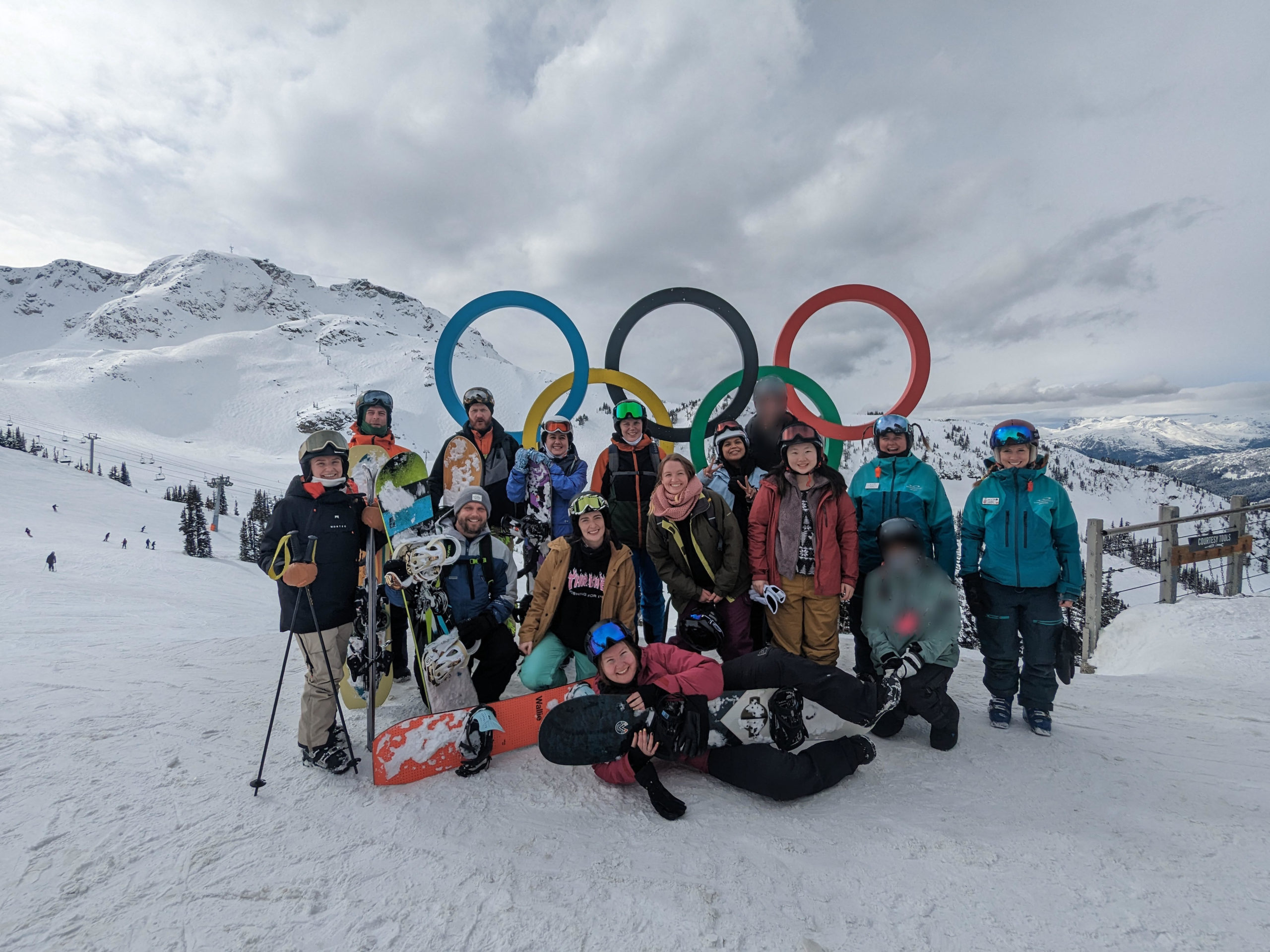 [Photo description: A group of 15 happy young adults -- 12 YACCers and three ski/snowboard instructors -- in winter wear and carrying skis/snowboards gather in front of the Olympic Rings with snowy mountains behind them.]